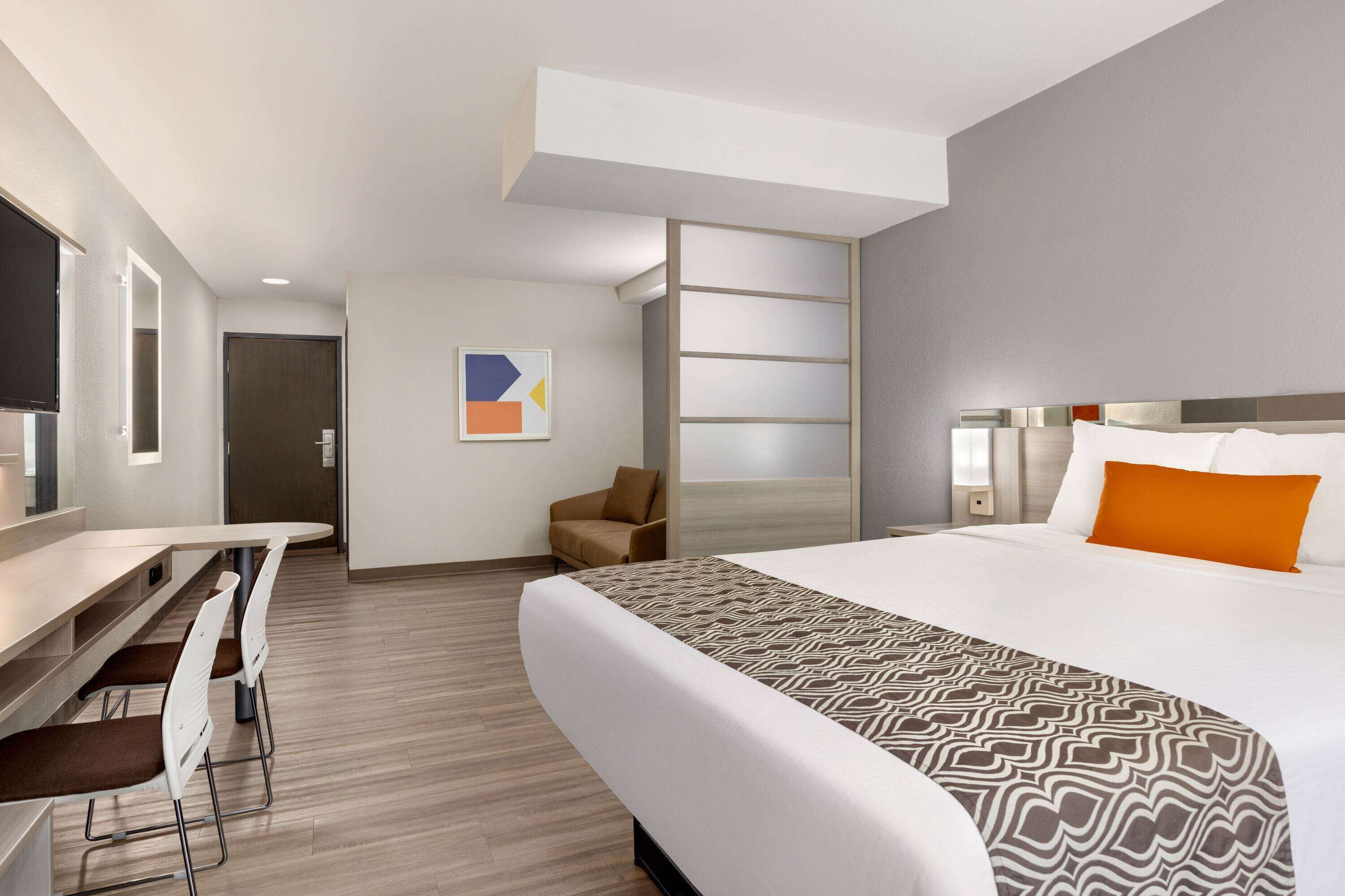 Una habitación de hotel moderna con una cama grande, un escritorio con sillas, un pequeño rincón de descanso y un amplio ventanal.