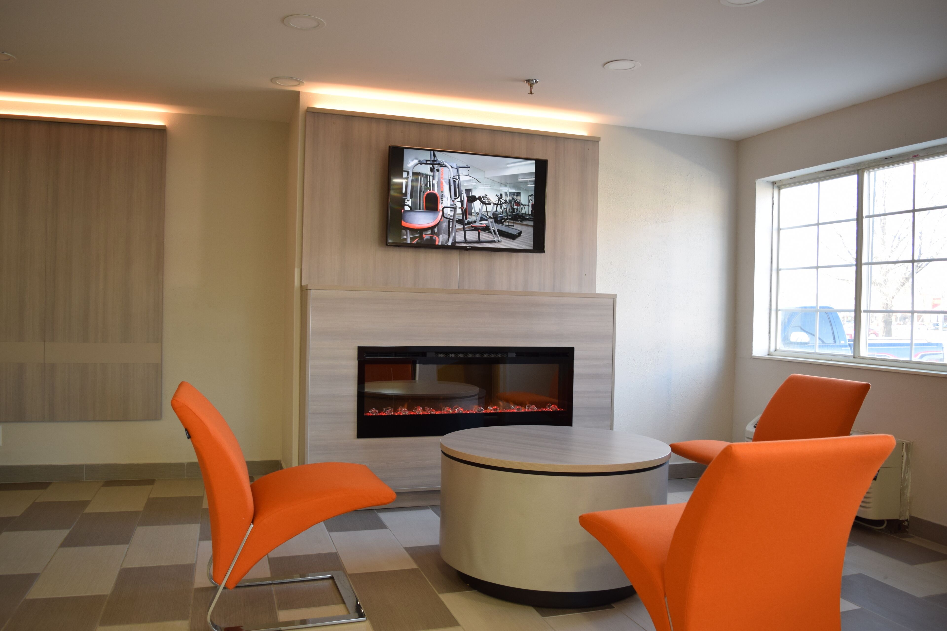 Una habitación moderna con televisor de pantalla plana sobre chimenea, sillones naranjas alrededor de una mesa redonda, y un ventanal que proporciona luz natural.