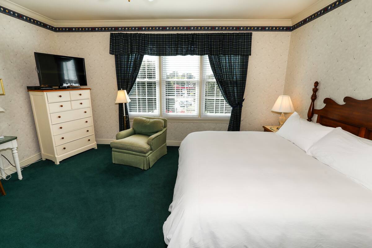 Ein Hotelzimmer mit Bett, Schminktisch, Fernseher, Sessel und einem Fenster mit Vorhängen.