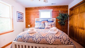 A bedroom with a wooden headboard, a bed with patterned bedding, a bedside table with a lamp, and a window with blinds. - Immaculate Family-Friendly Canal Front Vacation Home in Outer Banks 4x4 Area (Corolla)