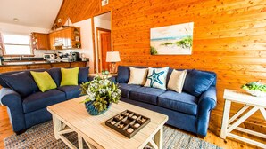 A cozy living room with a blue sofa, a wooden coffee table, and a painting of a beach scene. - Immaculate Family-Friendly Canal Front Vacation Home in Outer Banks 4x4 Area (Corolla)