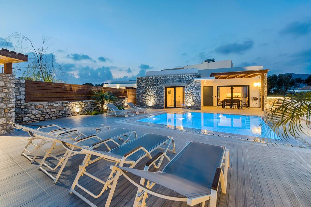 Un área de piscina moderna al aire libre con sillones de descanso, un muro de piedra y un edificio con puerta de vidrio.