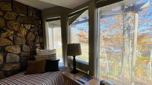 Une pièce chaleureuse avec un mur en pierre, deux lits avec une literie à rayures, une table de chevet en bois avec une lampe, et une grande fenêtre offrant une vue sur les arbres et un ciel dégagé.