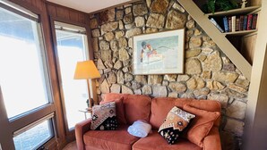 Un salon avec un mur en pierre, un canapé marron, des coussins à motifs, un pouf rond, une lampe et une bibliothèque.