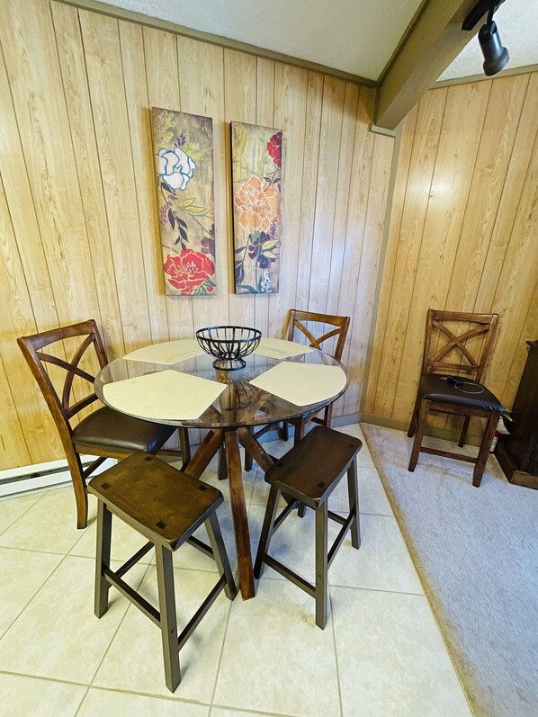 A dining area with a round glass table, six wooden chairs, and two floral paintings on the wall.