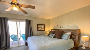 A bedroom with a bed, a ceiling fan, a nightstand, a lamp, a painting, and a view of the outside through a sliding door.