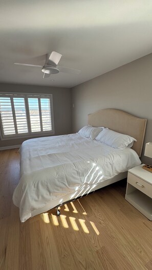 Une chambre à coucher comprenant un lit, une table de chevet, un ventilateur de plafond et une fenêtre avec des stores.