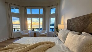 A hotel room with a large bed, a view of the sunset through the windows, and a seating area with chairs and a table.
