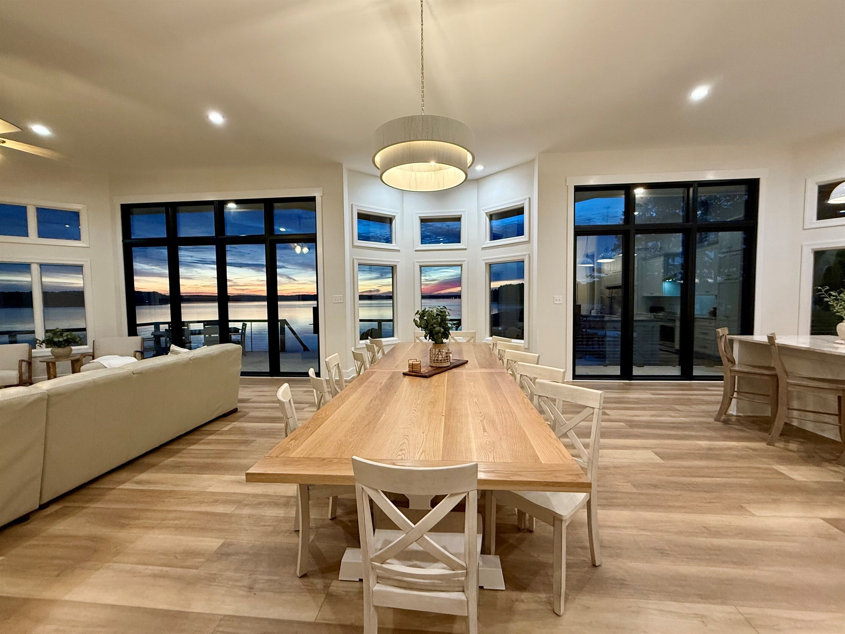 A spacious dining area with a long wooden table, white chairs, and large windows offering views of the sunset over a body of water.