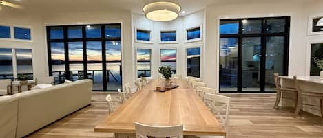 A spacious dining area with a long wooden table, white chairs, and large windows offering views of the sunset over a body of water.