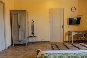 Ein Hotelzimmer mit Bett, Schreibtisch mit Stuhl, Kleiderschrank, Zimmerpflanze, Fernseher und einer Uhr an der Wand.