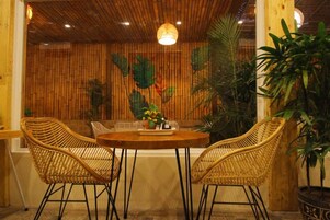 Una habitación con una mesa redonda de madera, dos sillas de mimbre y una planta grande en maceta.