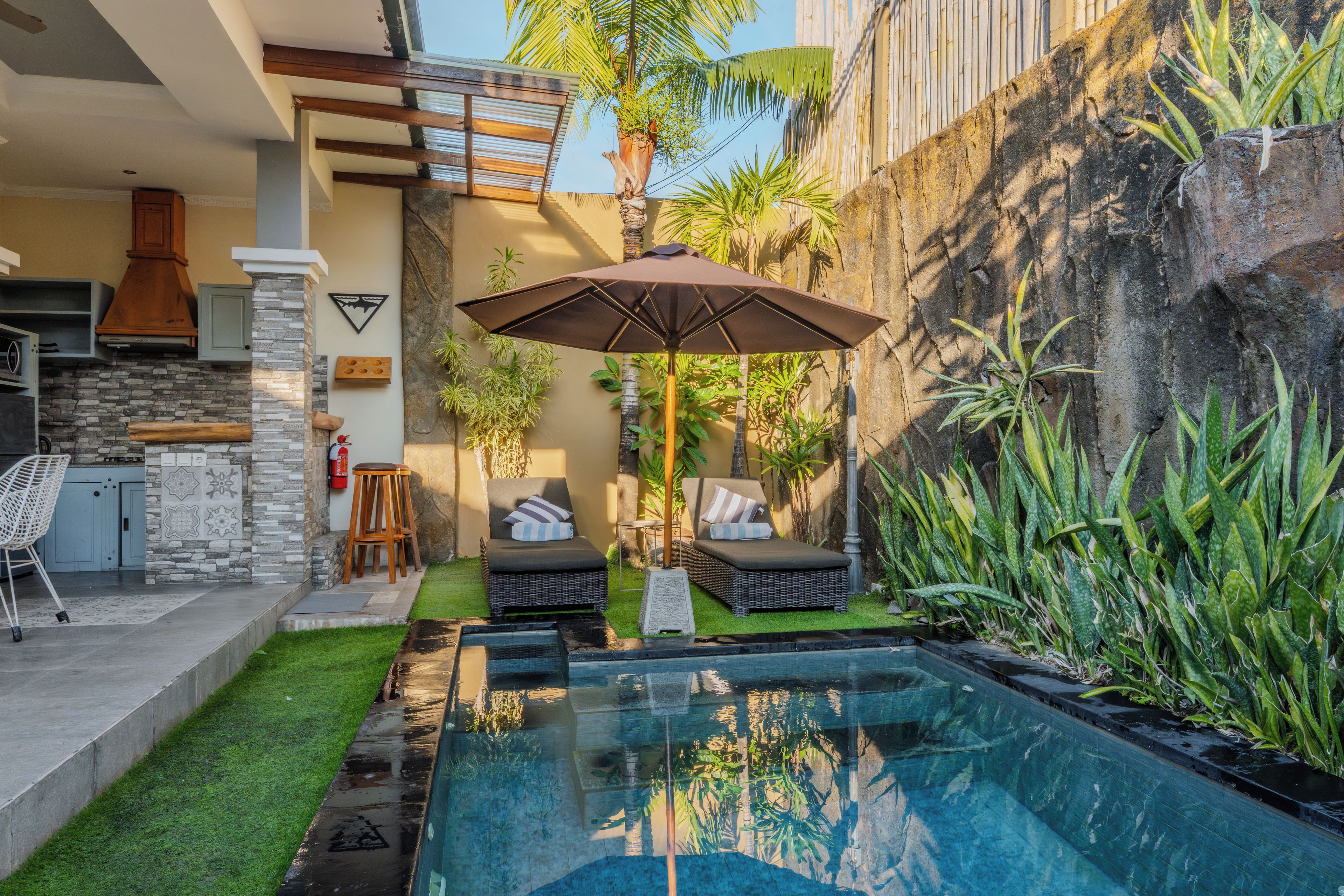 A pool area with two lounge chairs and a table, surrounded by tropical plants and a stone wall.