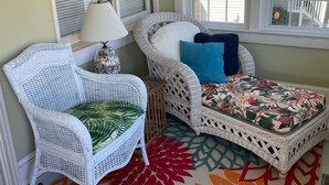 A room with a white wicker chair, a small table with a lamp, and a bench with a floral cushion. - Cottage On Famous Chincoteague Island-Famous Wild Ponies (Chincoteague Island)