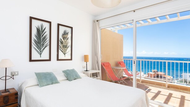 A bedroom with a large bed, two framed leaf artworks, a nightstand, a lamp, and a balcony with red chairs and a table overlooking the sea. - Beautiful sea view duplex - PORÍS 1 (Poris de Abona)