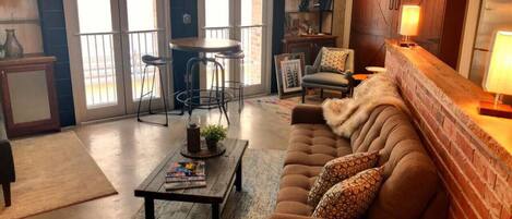 A cozy living room with a brown sofa, a coffee table, and a fireplace. There is a dining area with a table and chairs, and a kitchen area with cabinets and a refrigerator.