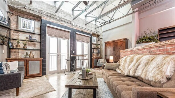 Un vaste espace de vie de style loft, avec des murs en briques apparentes, un plafond haut doté de fenêtres de toit, un grand canapé agrémenté d’un plaid en fourrure et une table basse rustique.