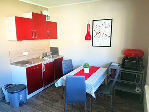 Cocina compacta con armarios rojos, un lavamanos, una pequeña mesa de comedor con sillas, una planta en maceta y un microondas.