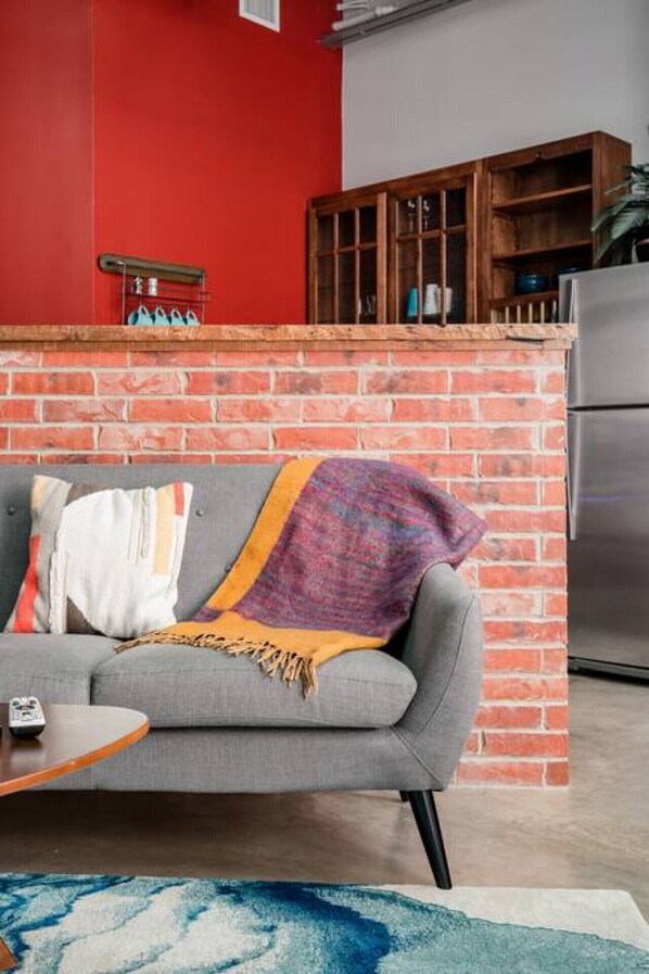 Ein Wohnzimmer mit grauem Sofa, einer roten Backsteinwand, einem Holzschrank und einem Edelstahlkühlschrank.