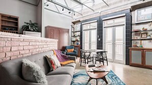 A modern living room with a grey sofa, a coffee table, and a blue patterned rug. There is a brick wall, a wooden cabinet, and a large window with a view. - Green Door Lofts-Retro Loft - Silos/Downtown (Waco)