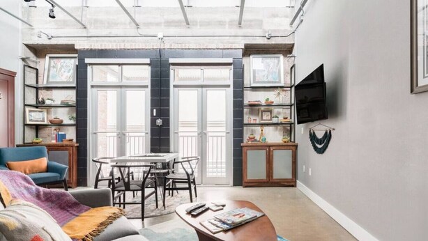 A spacious living area with a high ceiling, a dining table with chairs, a sofa, a coffee table, and a TV mounted on the wall. - Green Door Lofts-Retro Loft - Silos/Downtown (Waco)