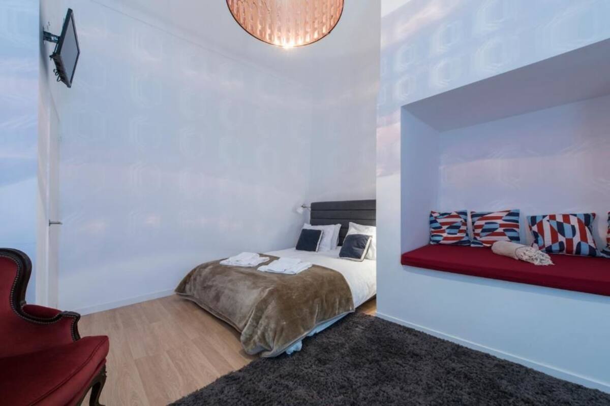 Un dormitorio con una cama, una silla roja, un banco empotrado con cojines estampados y una lámpara colgante.