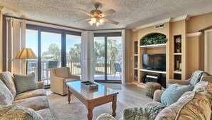 A spacious living room with a ceiling fan, large windows, a flat-screen TV, and a comfortable seating area. - Litchfield Beach 3Bed/3Bath w Sleeper sofa Oceanfront Condo in Pawleys Island SC (Pawleys Island)