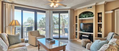 A spacious living room with a ceiling fan, large windows, a flat-screen TV, and a comfortable seating area.