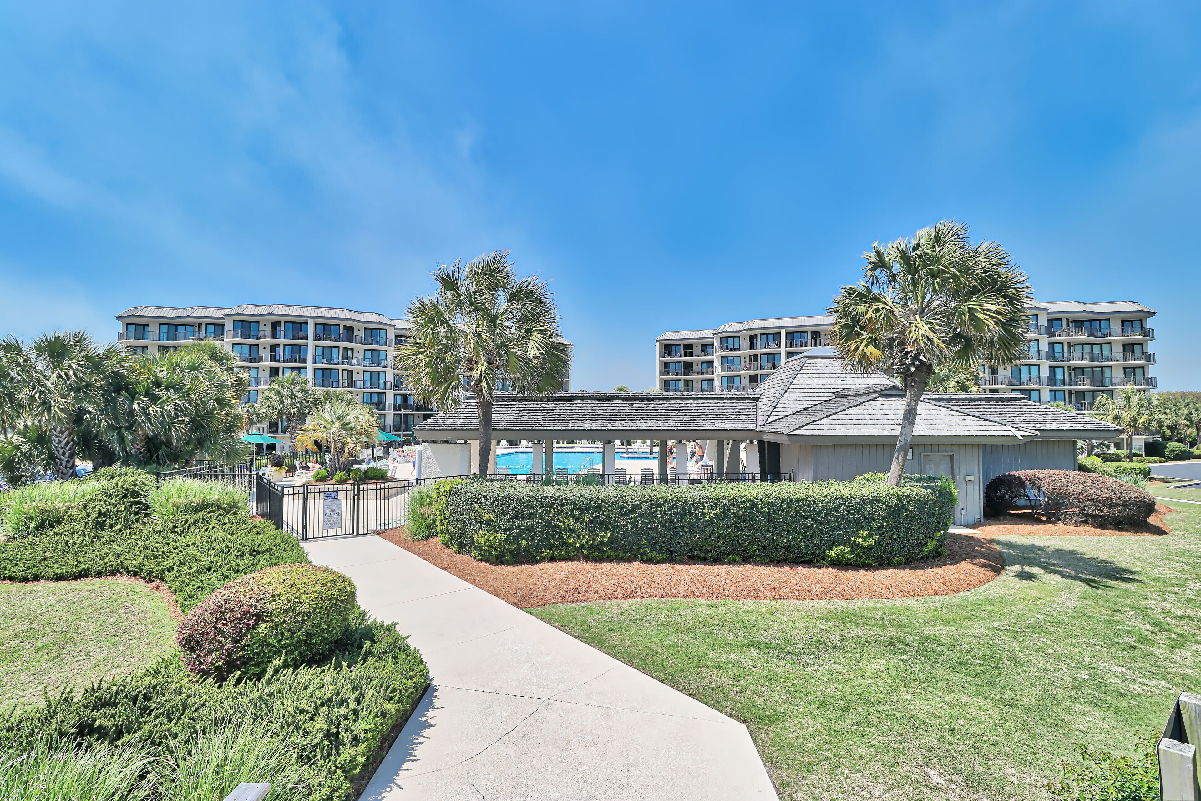 A modern hotel with a well-maintained lawn, a pathway leading to the entrance, and a pool area visible in the background.
