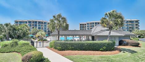 A modern hotel with a well-maintained lawn, a pathway leading to the entrance, and a pool area visible in the background.