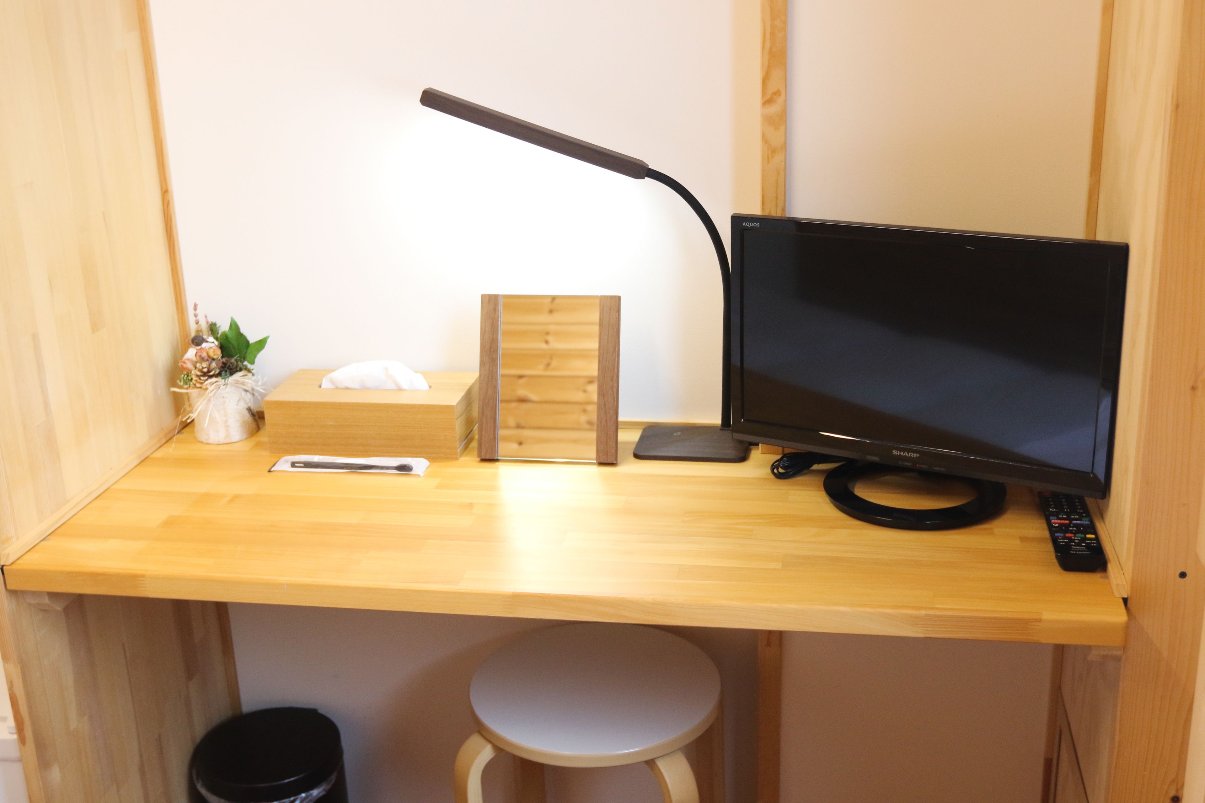 A wooden desk with a monitor, a lamp, a remote control, a small stool, and a trash bin.