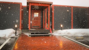 A building entrance with a red door and steps leading up to it. Snow is falling, and the ground is wet. - Phottage inn Biei (Biei)