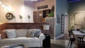 A cozy living room with a white sofa, a small side table, and a wicker chair. A sign reading "GROCERIES" is visible on the wall. - Green Door Lofts -Magnolia Loft, Silos/Downtown (Waco)