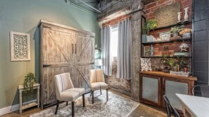A room with a large wooden wardrobe, two upholstered chairs, a patterned rug, a fireplace with decorative items, and a brick wall. - Green Door Lofts -Magnolia Loft, Silos/Downtown (Waco)