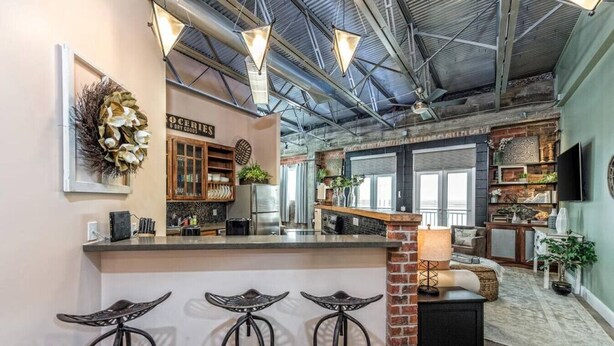 A spacious kitchen with a bar area, featuring a brick accent wall, pendant lighting, and a view into the living area. - Green Door Lofts -Magnolia Loft, Silos/Downtown (Waco)