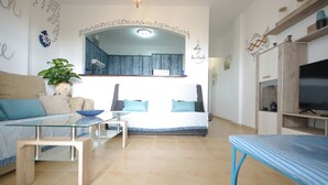 Un soggiorno con un divano, un tavolino in vetro, una televisione e un angolo cottura sullo sfondo.