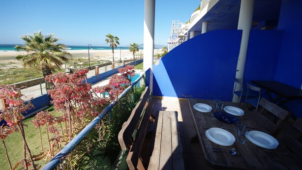Un balcone con tavolo in legno apparecchiato per mangiare, con vista su una spiaggia, palme e cielo sereno.