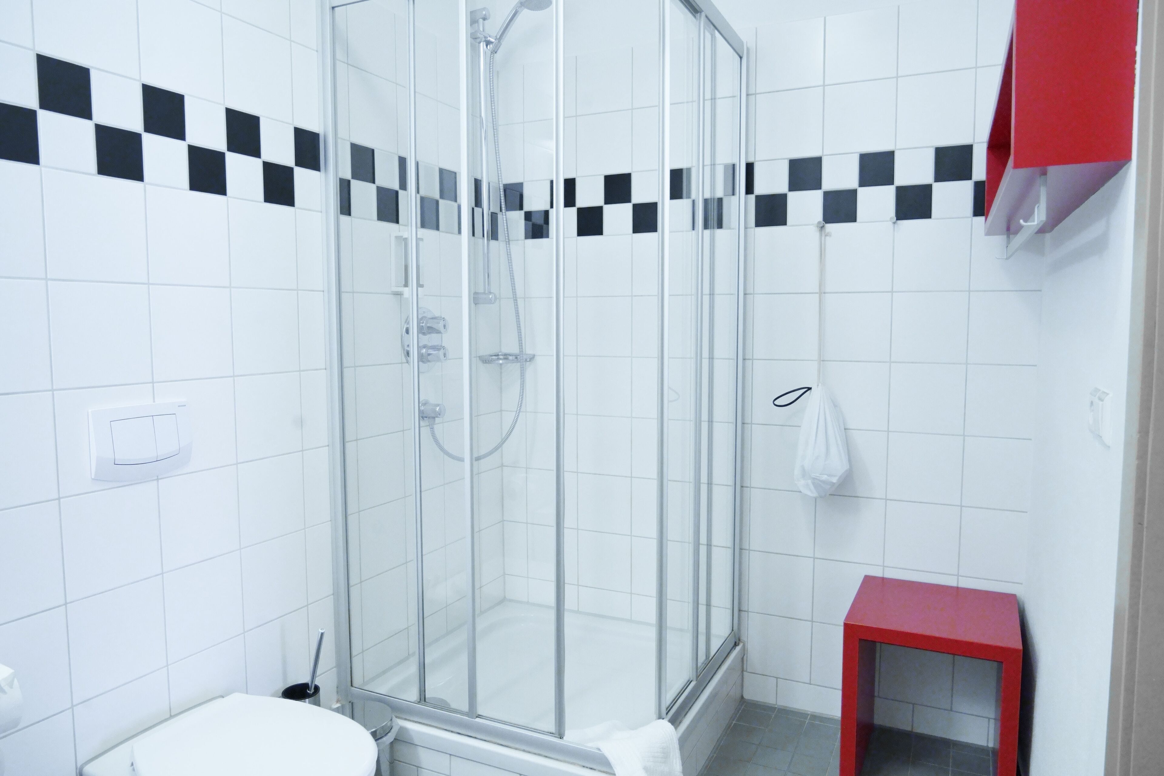 A modern bathroom with a glass-enclosed shower, white tiled walls, a red stool, and a checkered black and white tile border.