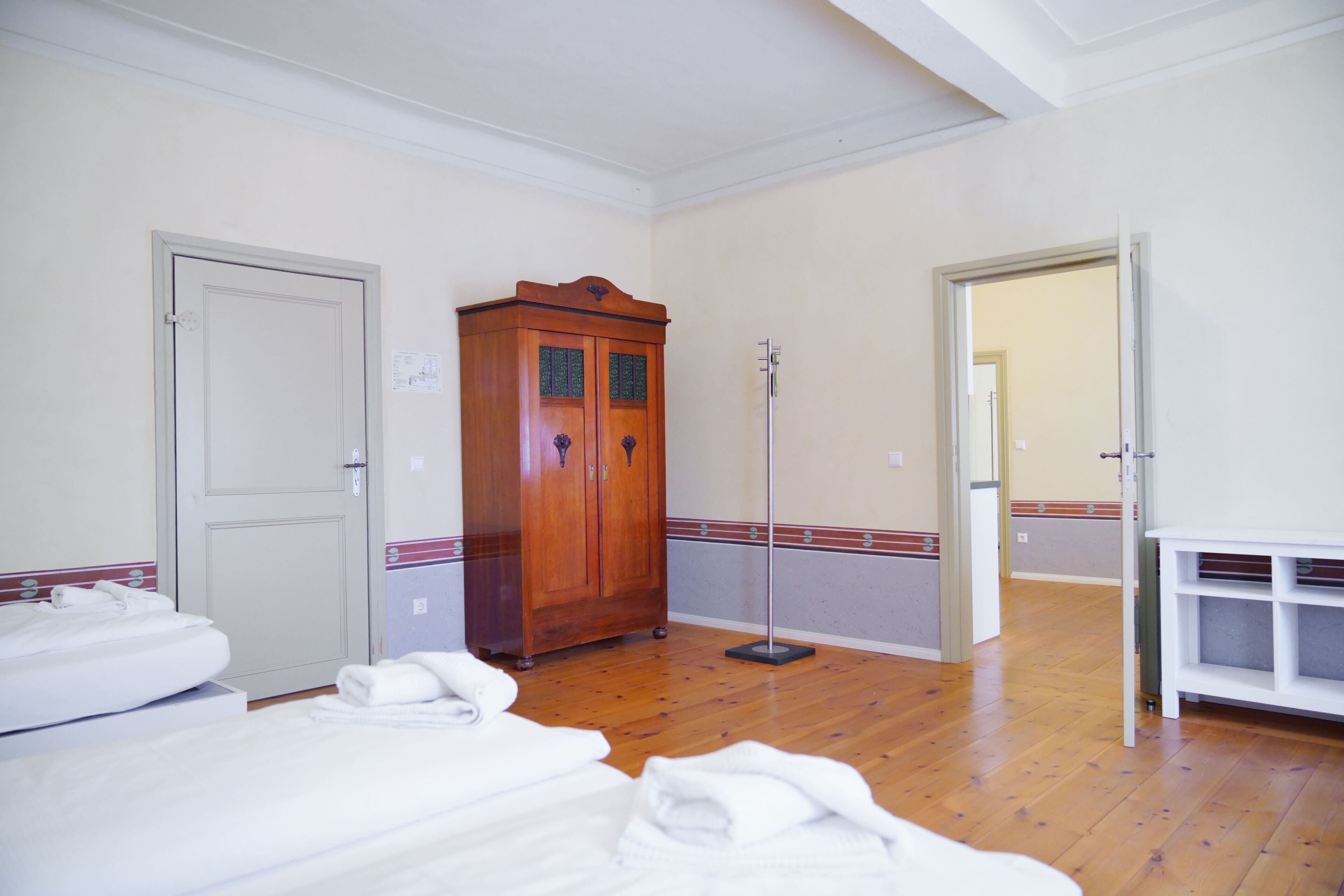 Quarto de hotel com soalho de madeira, guarda-roupa, candeeiro de pé e cama com roupa de cama branca.