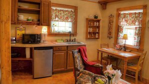 A cozy kitchen with wooden cabinets, a microwave, a sink, and a dining area with a chair and table. - Rumpelstilzchen Cottage (#2) (Fredericksburg)