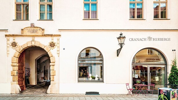 A building with a stone archway entrance, a storefront with a sign reading "CRANACH HERBERGE," and outdoor seating with a bicycle parked outside. - Cranach-Grand - Cranach-Herberge Wittenberg (Lutherstadt Wittenberg)