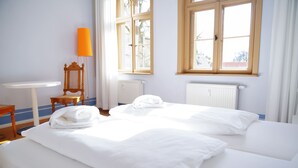 Quarto de hotel com duas camas, uma mesinha, uma cadeira e uma janela com cortinas.