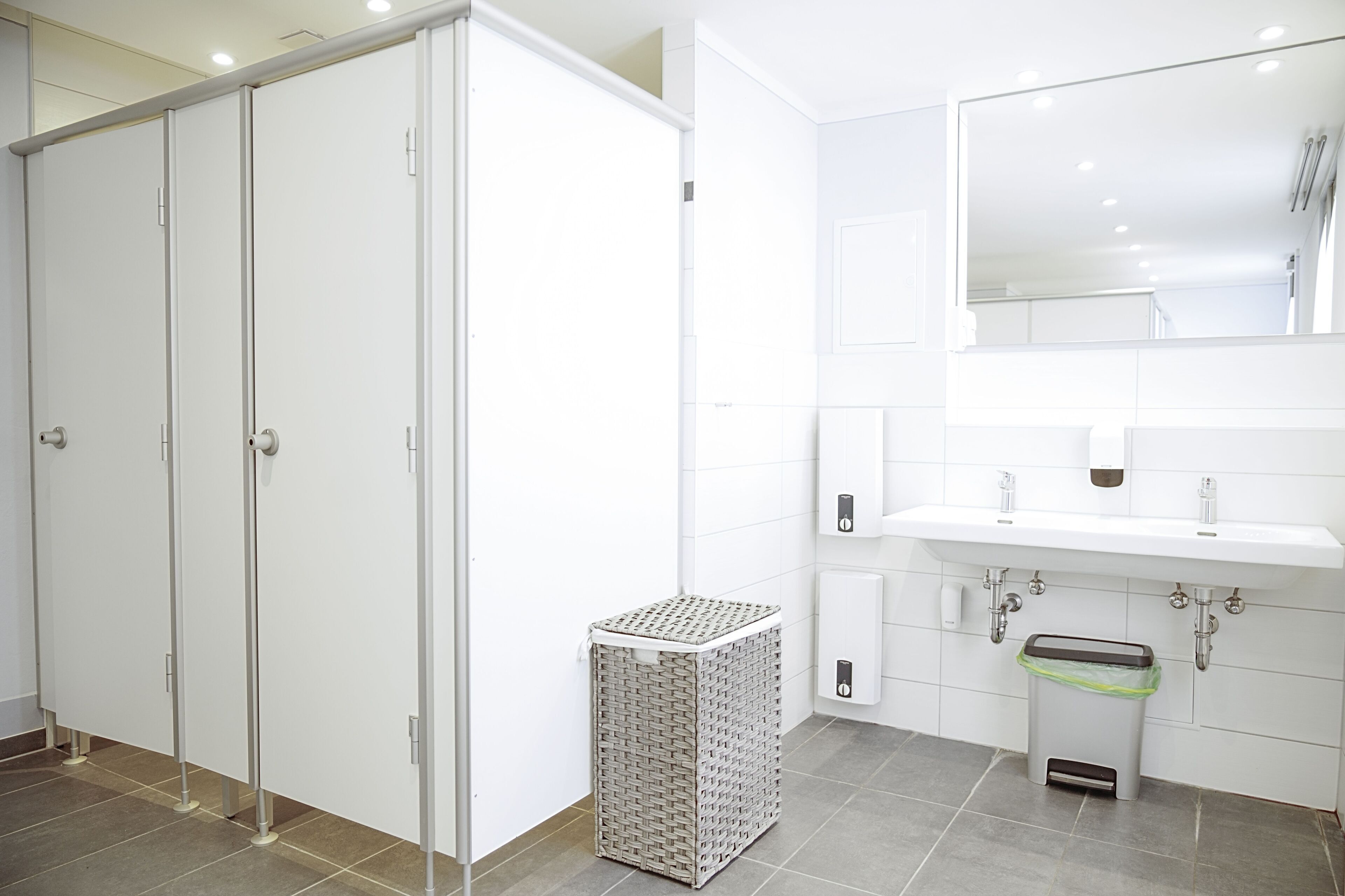 Ein modernes Badezimmer mit Waschbecken, Spiegel und Fliesenboden. Weiße Schränke und ein Mülleimer sind ebenfalls vorhanden.