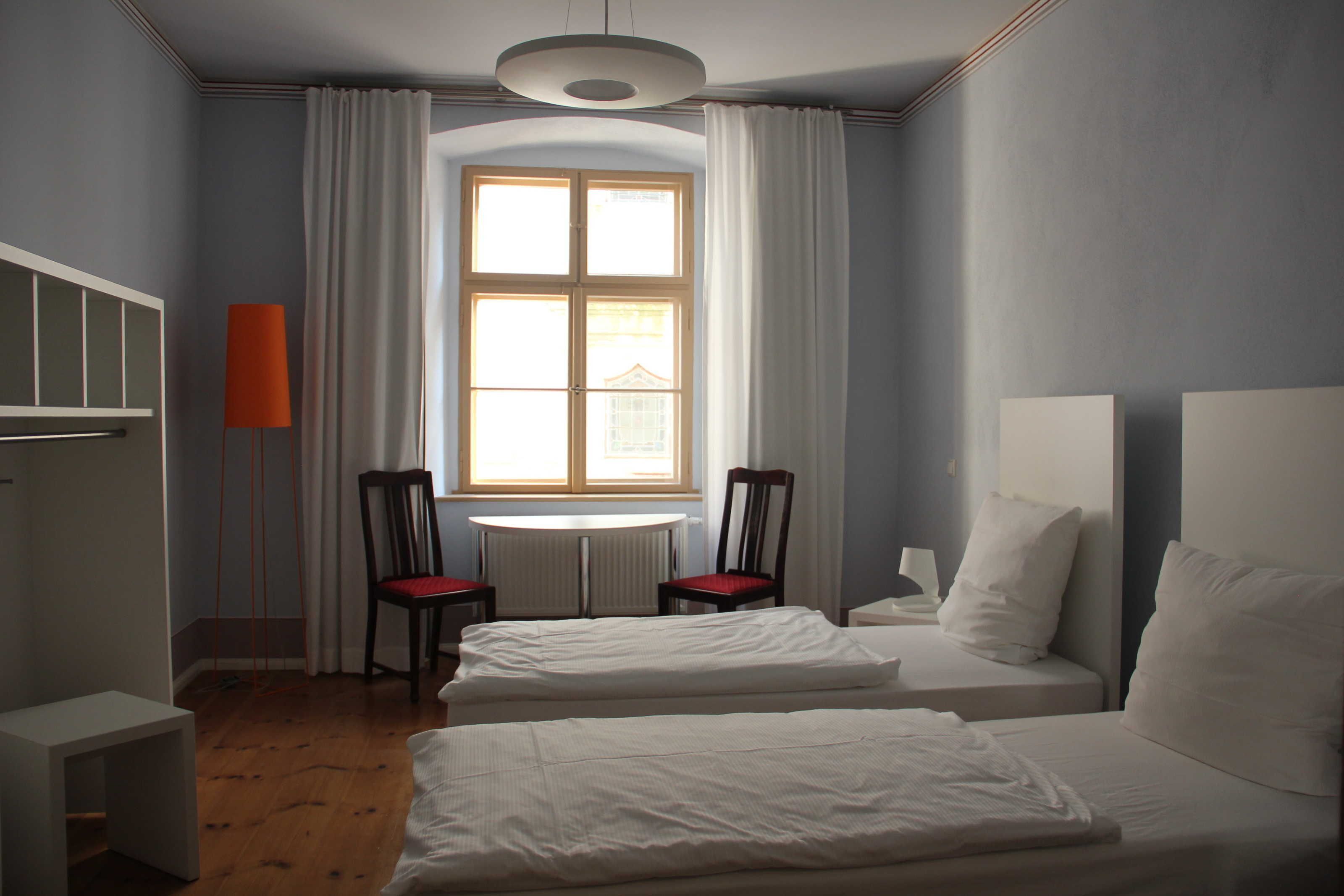 A hotel room with two beds, a round table, two chairs, a radiator, and a window with curtains.