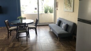 Un espace de vie compact comprenant une table et des chaises, un canapé, un réfrigérateur et une grande fenêtre offrant une vue sur l’extérieur.