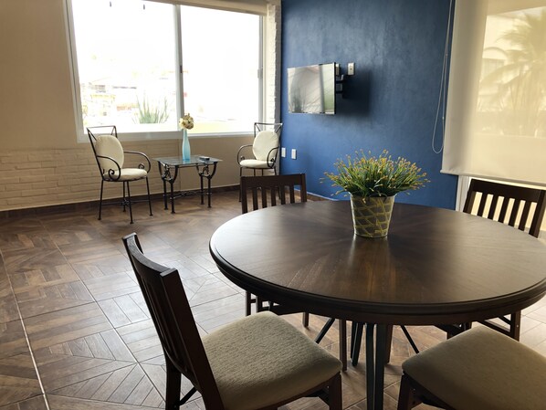 Ein Konferenzraum mit einem runden Tisch, Stühlen, einem Blumentopf, einem an der Wand montierten Fernseher und einem Fenster mit Außenblick.