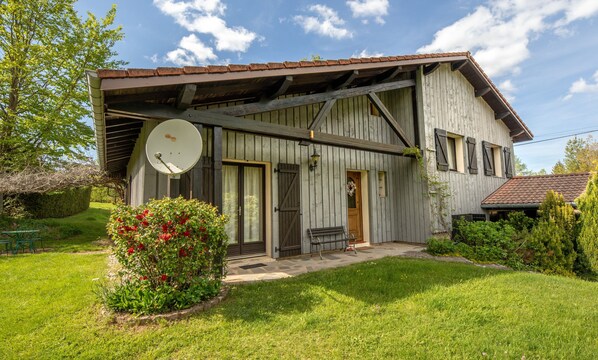 Une maison en bois rustique, dotée d’une antenne parabolique, d’un banc et d’un jardin où poussent des fleurs rouges.