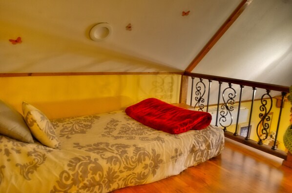Un dormitorio con cama, almohadas, una colcha roja, suelo de madera, techo blanco y barandilla de balcón.
