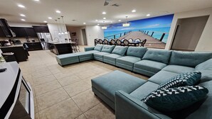 A modern living room with a large L-shaped sectional sofa, a dining area with a wooden table and chairs, and a kitchen with dark cabinets and stainless steel appliances. - Spacious Villa for Large Groups ✦ Lazy River and Tiki Hut ✦ (Kissimmee)
