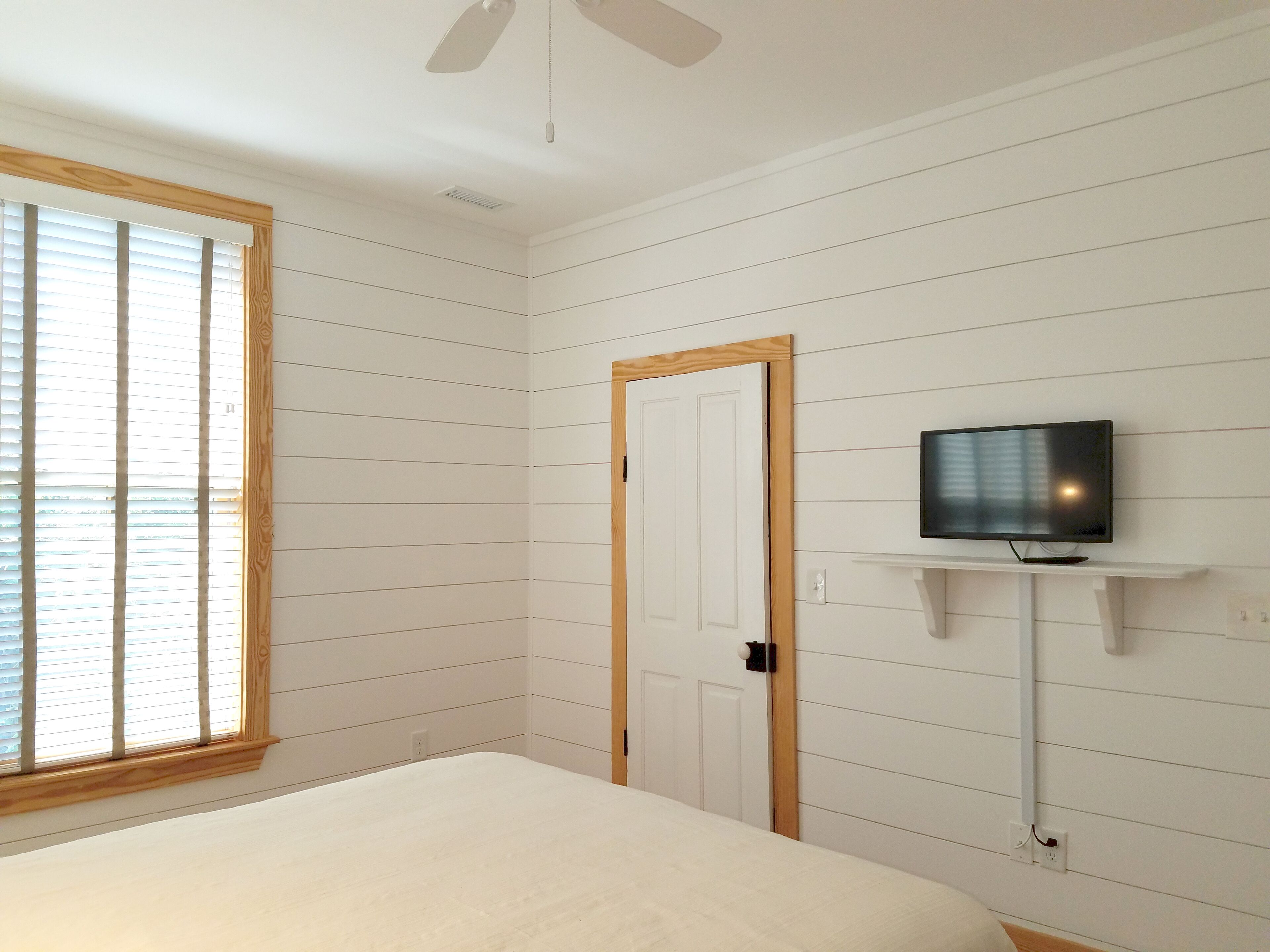 Une chambre à coucher avec un lit, un téléviseur posé sur un meuble, une porte et une fenêtre avec des stores.